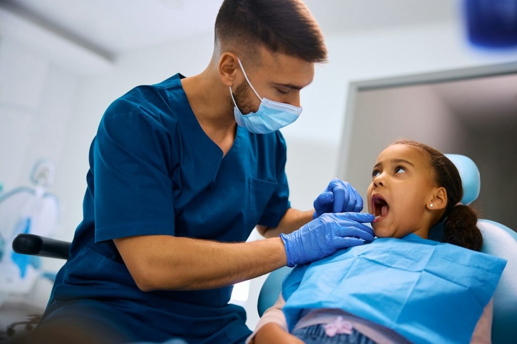 Male dentist examining teeth of black little girl at dental clinic.