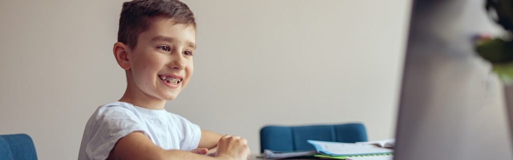 Portrait of cute small happy boy with braces having online video lesson at laptop.